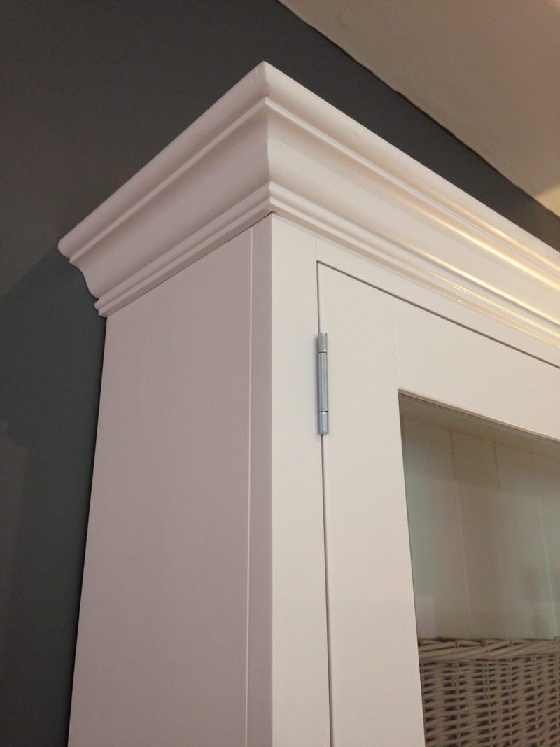 Close view of the top corner of the painted dresser top, showing the deep moulded cornice, the flat side panel and a brushed steel hinge on the glazed door frame, set against a dark grey wall