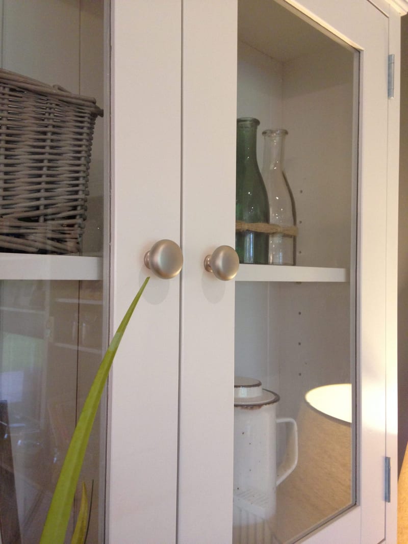 Close-up of the pair of satin nickel knobs on the glazed doors of the dresser top, with a grey wicker basket, a green bottle, a clear bottle and a white enamel jug on the shelves behind