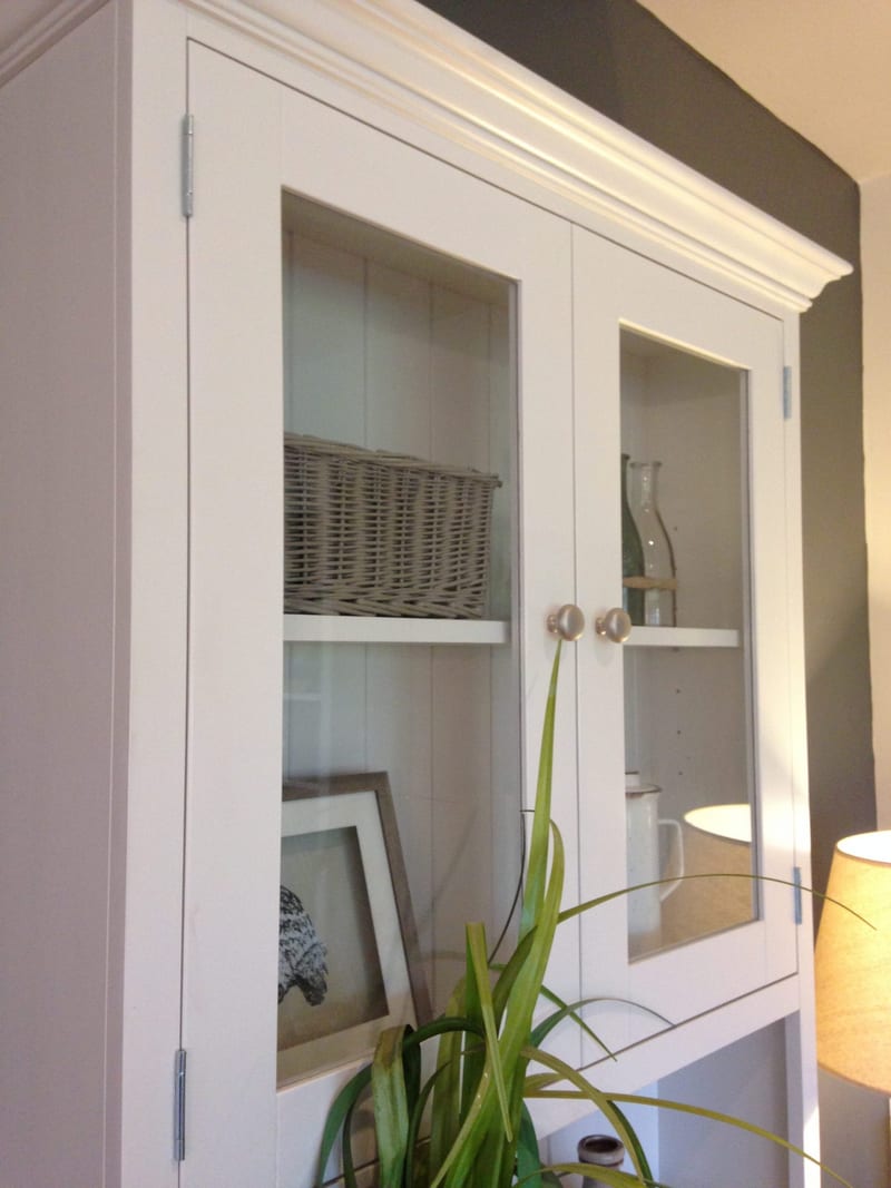 Angled view of the glazed dresser top from the left, showing the moulded cornice, tongue-and-groove back boards, a wicker basket on the shelf and a framed print leaning below