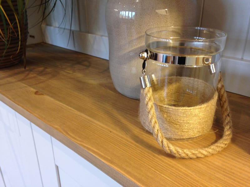 Close view along the waxed solid wood worktop, showing the knot and grain of the timber beside a glass storage jar with a rope handle and a large grey ceramic bottle