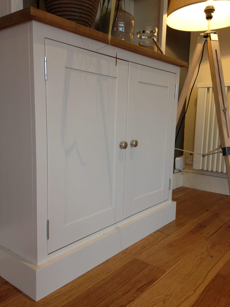 Angled view of the base cupboard from the left, showing its two panelled doors, a pair of round satin knobs, the waxed timber top above and the plinth on an oak plank floor