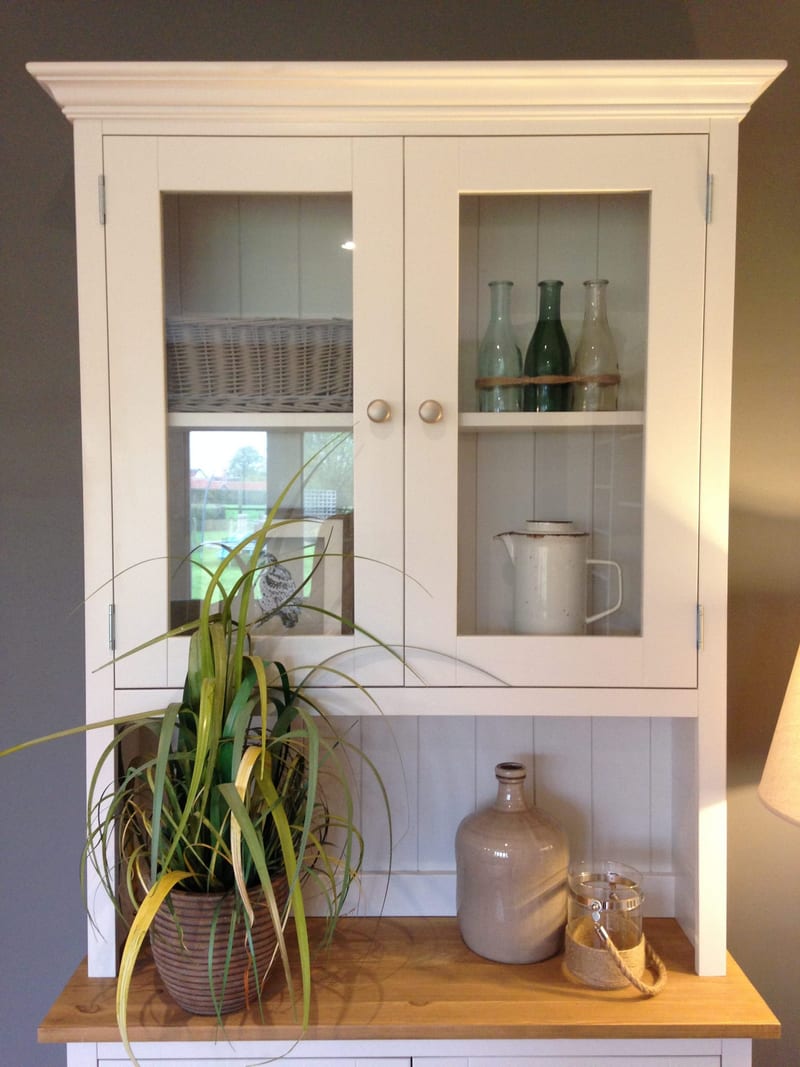 Full view of the glazed dresser top with its moulded cornice, two glass-panelled doors and satin knobs, holding a wicker basket, green bottles and an enamel jug above a grassy plant