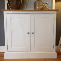 Front view of the painted base cupboard with two panelled doors, two round satin knobs and a waxed timber top, dressed with a potted plant, ceramic flagon and rope-handled lantern
