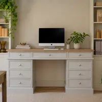 Front view of the grey painted desk between two tall cabinets with open shelves, showing its oak top dressed with an all-in-one computer, a brass bowl, stacked books and a leafy plant