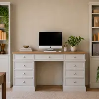 Wide view of the desk on a natural fibre rug between two tall painted cabinets with open shelves and panelled doors, set against a plain putty coloured wall with an upholstered chair in front