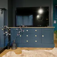 Front view of the navy painted media unit with an oak top, a cupboard at each end and three central drawers with brass knobs, a television above and blossom stems in a smoked glass vase beside