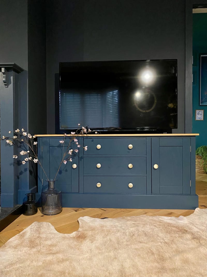 Front view of the navy painted media unit with an oak top, a cupboard at each end and three central drawers with brass knobs, a television above and blossom stems in a smoked glass vase beside