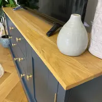 Close view along the oak top of the navy unit, showing the grain and squared edge beside a crackle glazed cream vase, with the brass knobs and the foot of the television alongside