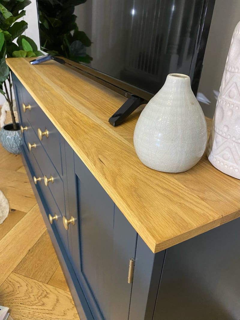 Close view along the oak top of the navy unit, showing the grain and squared edge beside a crackle glazed cream vase, with the brass knobs and the foot of the television alongside