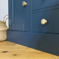 Low corner view of the end cupboard and two drawers, with turned brass knobs, two chrome hinges, the deep plinth and a woven seagrass basket beside on a herringbone oak floor