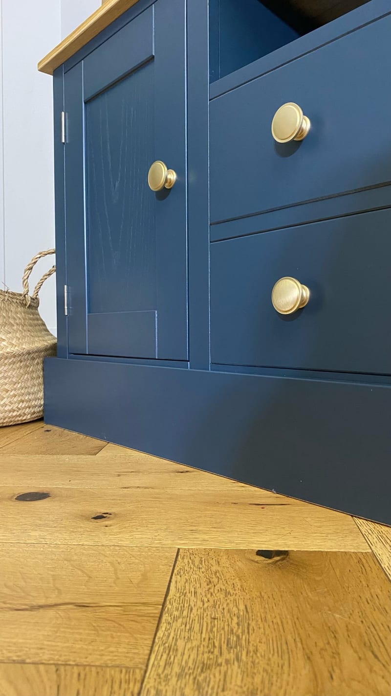 Low corner view of the end cupboard and two drawers, with turned brass knobs, two chrome hinges, the deep plinth and a woven seagrass basket beside on a herringbone oak floor