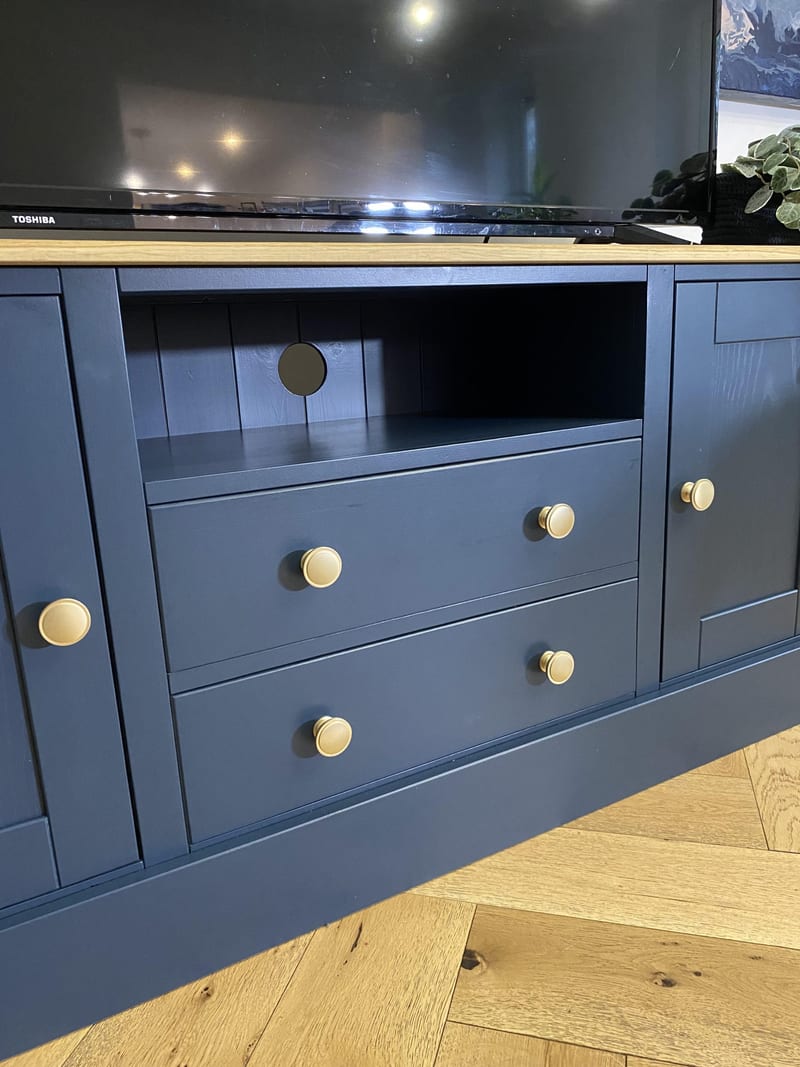 Close front view of the open media shelf with its round cable port and tongue-and-groove back, above two brass knobbed drawers and between the two end cupboards, with the television above