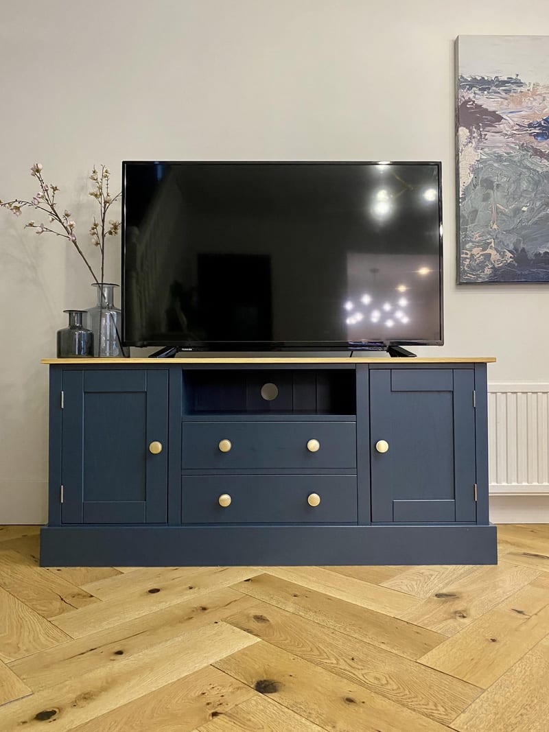 Full front view of the navy media unit in a pale room, with two panelled end cupboards, an open shelf, two drawers, brass knobs and an oak top, on a herringbone oak floor beside a radiator