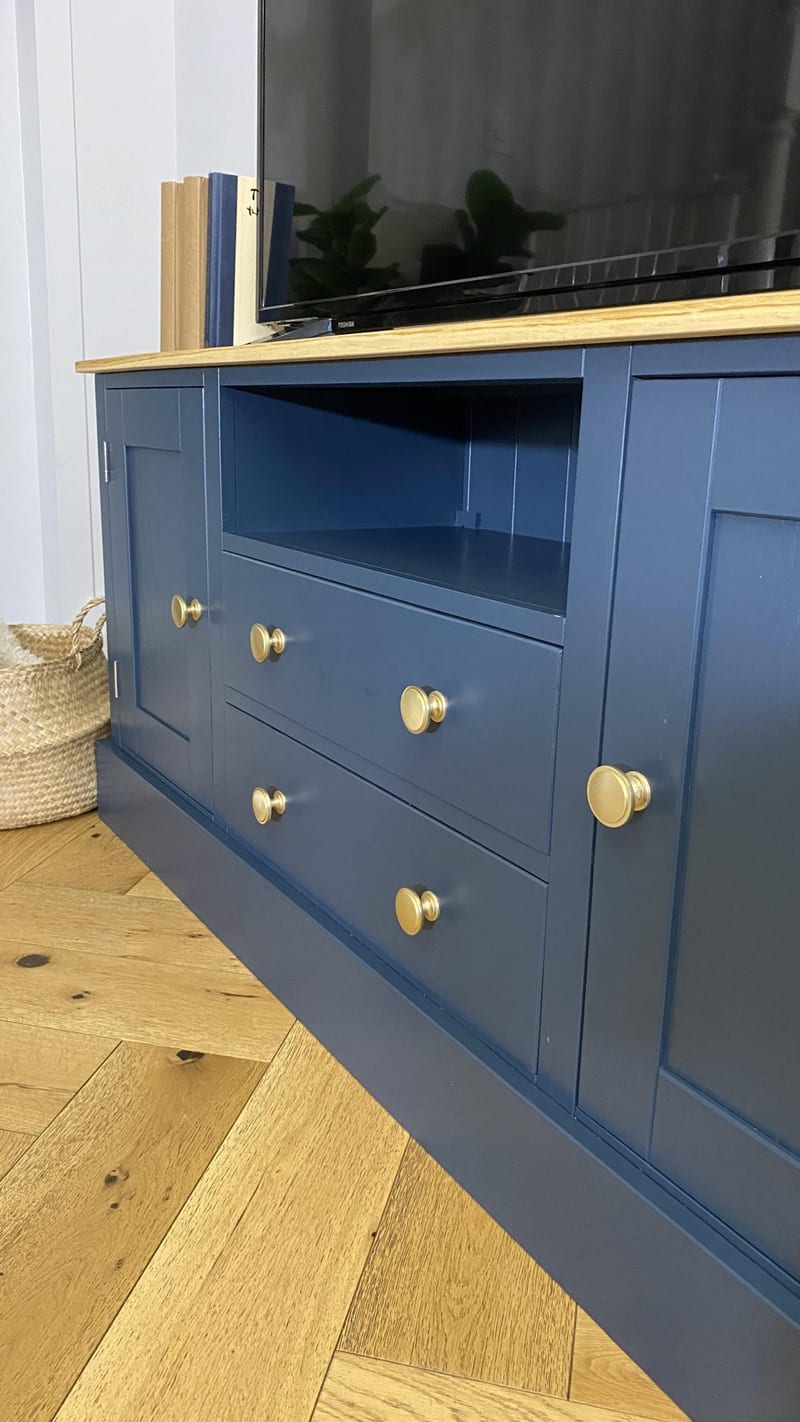Angled view along the unit from the right, showing the panelled cupboard door nearest the camera, two brass knobbed drawers, the open media shelf and a seagrass basket on the herringbone floor