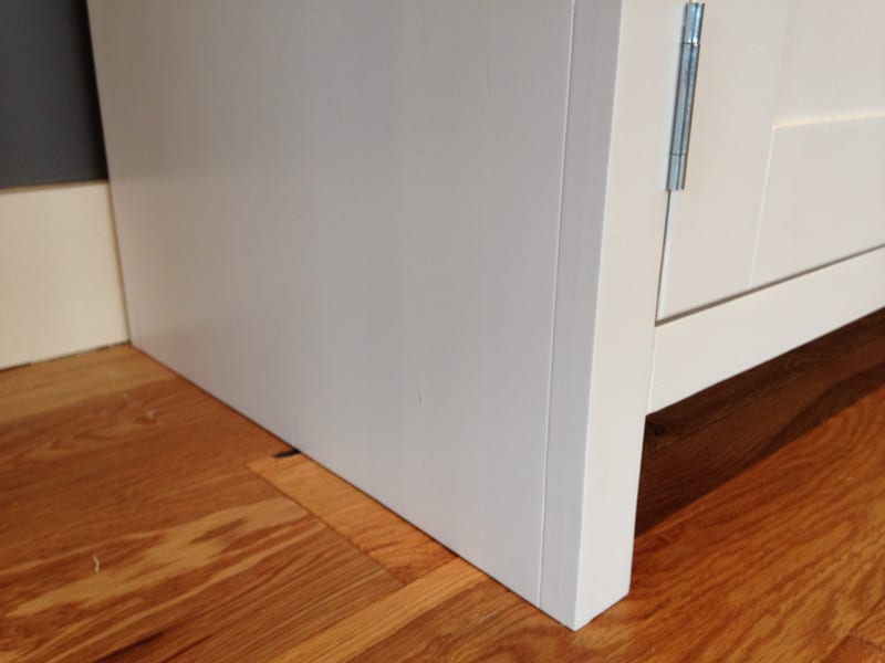 Close view of the lower corner of the painted sideboard, showing the plain end panel, a chrome hinge on the neighbouring shaker door and oak floorboards against a cream skirting board