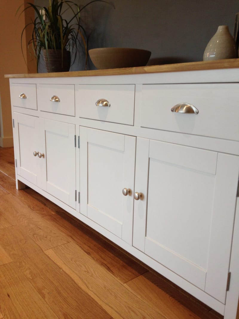 Angled close view of the white painted sideboard, showing four drawers with brushed nickel cup handles above four shaker doors with round knobs, and the natural wood top on an oak floor