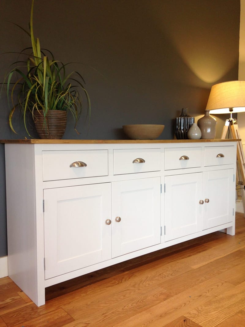 Full-length angled view of the white sideboard against a dark grey wall, four cup-handled drawers over four shaker doors, with a potted plant, stone bowl and vases on the wooden top