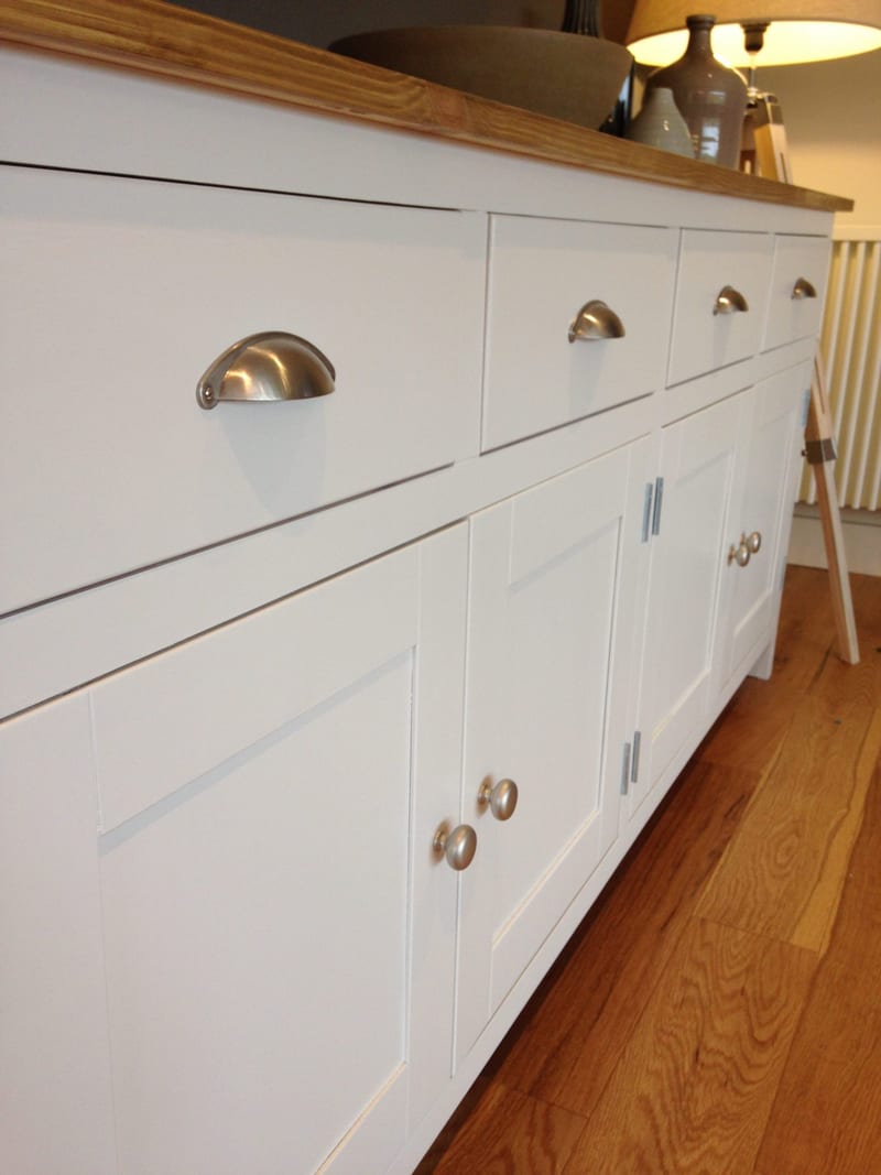 Low angled view along the front of the sideboard, showing the brushed nickel cup handles catching the light, the shaker door fronts with round knobs and the overhanging wooden top