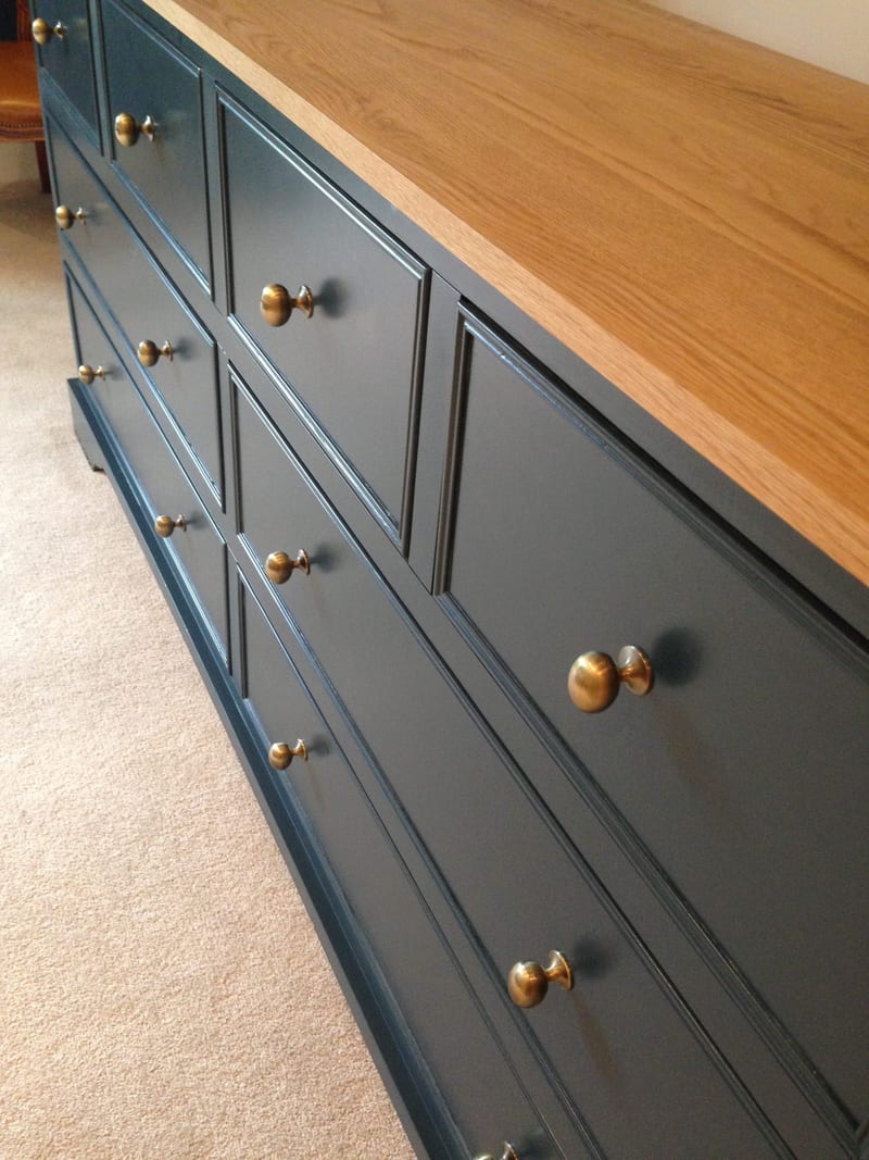 Angled close view along the dark blue chest of eight drawers, showing the beaded drawer fronts, aged brass knobs and the light oak top running to the far end above cream carpet