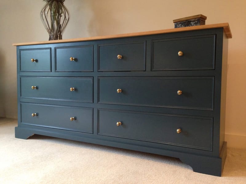Three-quarter view of the dark teal-blue eight-drawer sideboard with oak top, brass knobs and shaped bracket plinth, styled with a wire vase and small trinket box against a cream wall