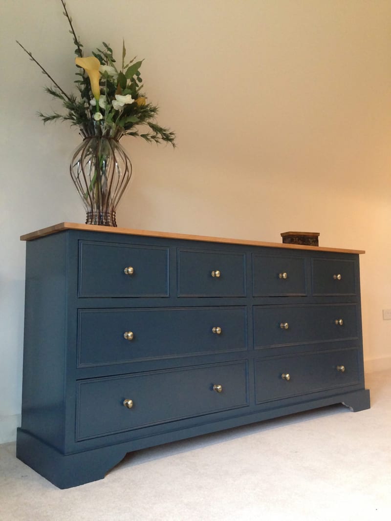 Angled view of the navy painted sideboard showing four short drawers above two rows of wide drawers, aged brass knobs, an oak top and a glass vase of yellow calla lilies and foliage