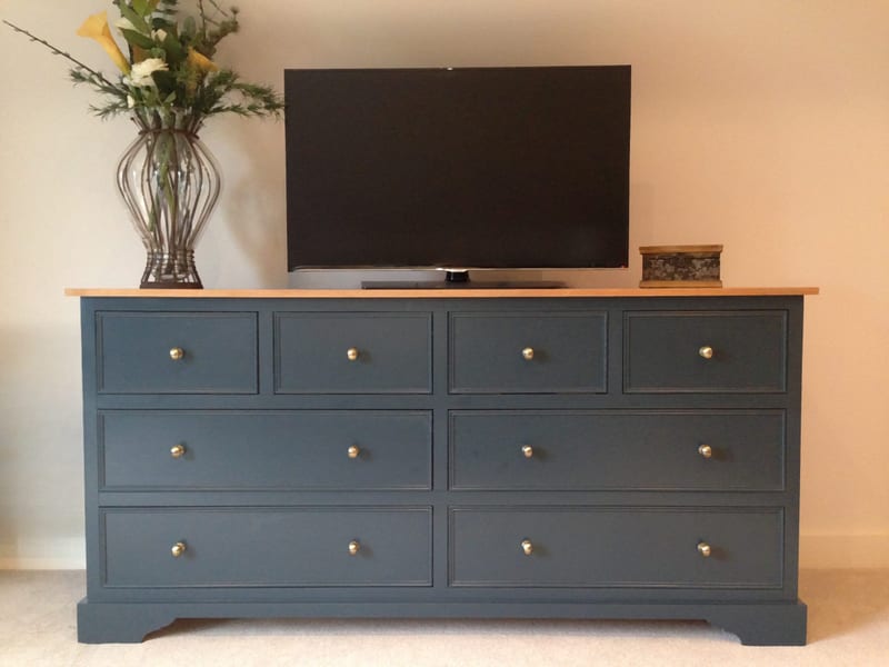 Front view of the dark blue eight-drawer sideboard used as a television stand, with a flat-screen TV, a vase of calla lilies and a small wooden box on the light oak top