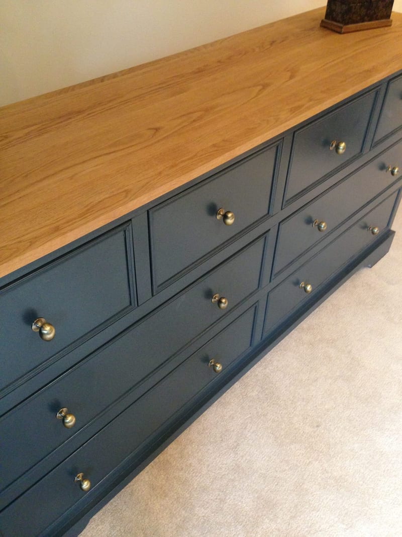 Close angled view of the light oak top of the navy sideboard, showing the open grain and square edge above the beaded drawer fronts and their aged brass knobs, with cream carpet below