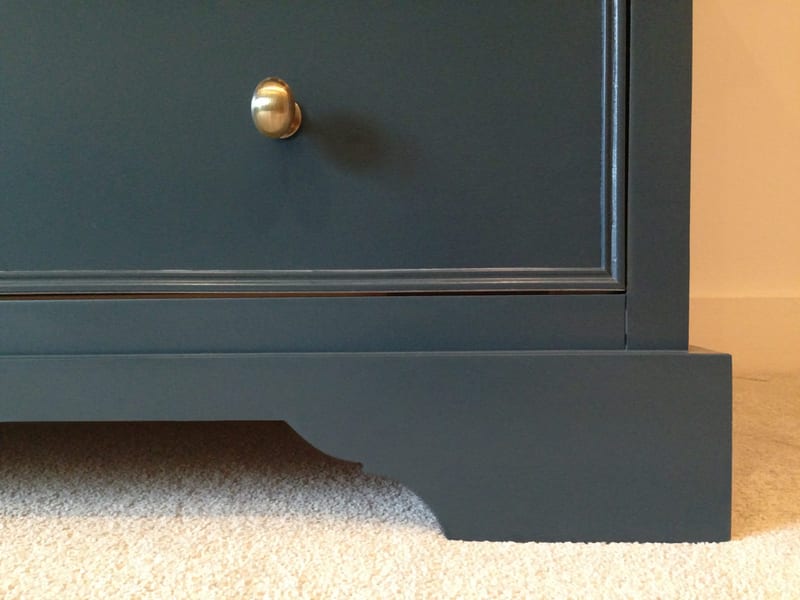 Close-up of the lower corner of the navy painted sideboard, showing a brass knob, the moulded drawer edge and the shaped bracket foot of the plinth resting on cream carpet