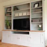 Angled full view of the grey painted media unit with three open shelved bays over an oak top, four shaker doors, an open television recess and a screen, on wide oak floorboards