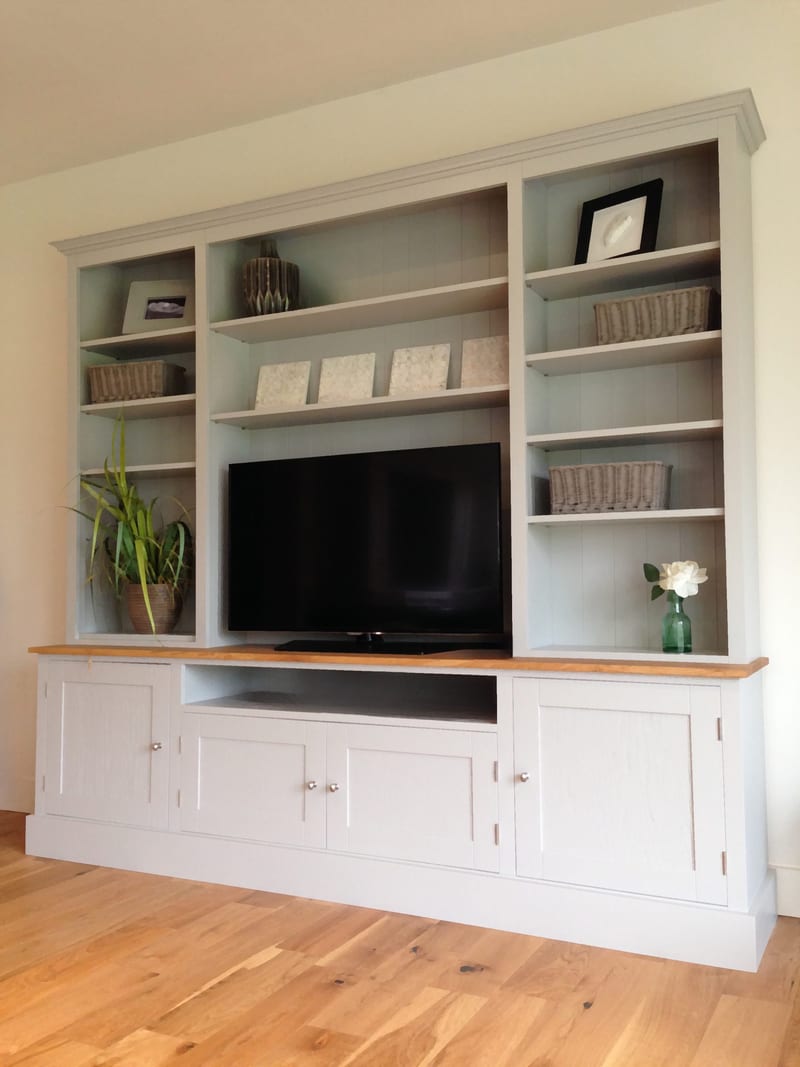 Angled full view of the grey painted media unit with three open shelved bays over an oak top, four shaker doors, an open television recess and a screen, on wide oak floorboards