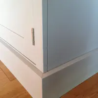 Close view of the lower corner of the grey painted unit, showing the plain side panel, a chrome hinge on the door edge and the stepped plinth meeting the oak floorboards