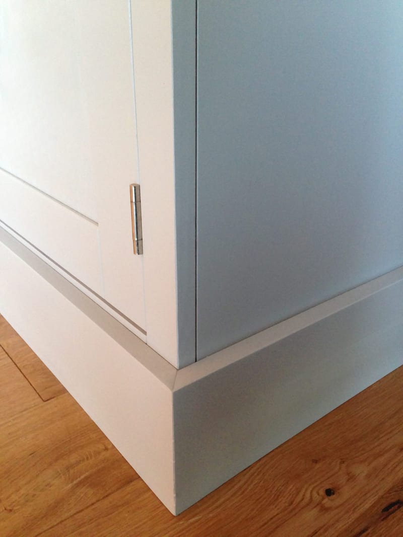 Close view of the lower corner of the grey painted unit, showing the plain side panel, a chrome hinge on the door edge and the stepped plinth meeting the oak floorboards