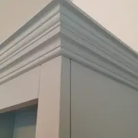 Close detail of the top corner of the painted media unit, showing the layered moulded cornice and the square corner post beneath, set against a pale wall and white ceiling