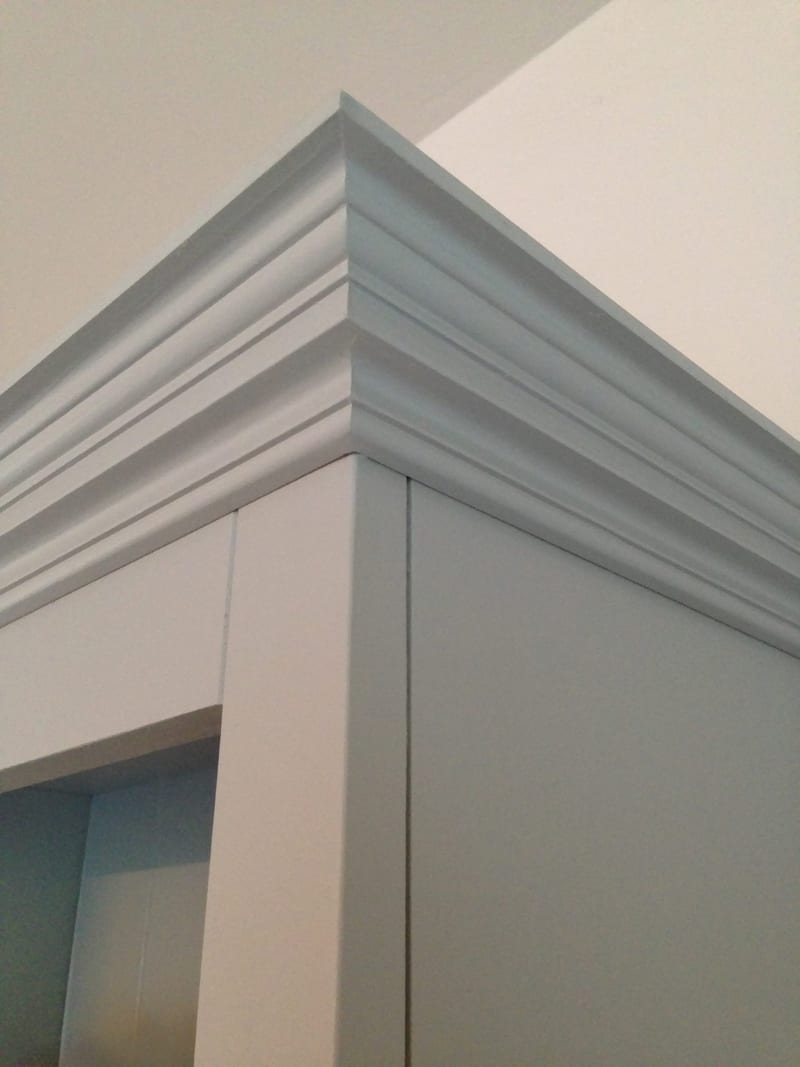 Close detail of the top corner of the painted media unit, showing the layered moulded cornice and the square corner post beneath, set against a pale wall and white ceiling