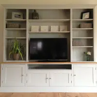 Straight-on view of the media unit with three shelved bays, pale grey-green tongue-and-groove backs, wicker baskets, two framed prints, a spider plant and a television above the cupboard base
