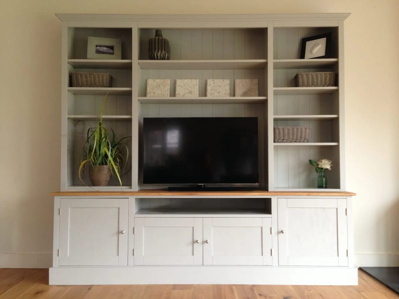 Straight-on view of the media unit with three shelved bays, pale grey-green tongue-and-groove backs, wicker baskets, two framed prints, a spider plant and a television above the cupboard base