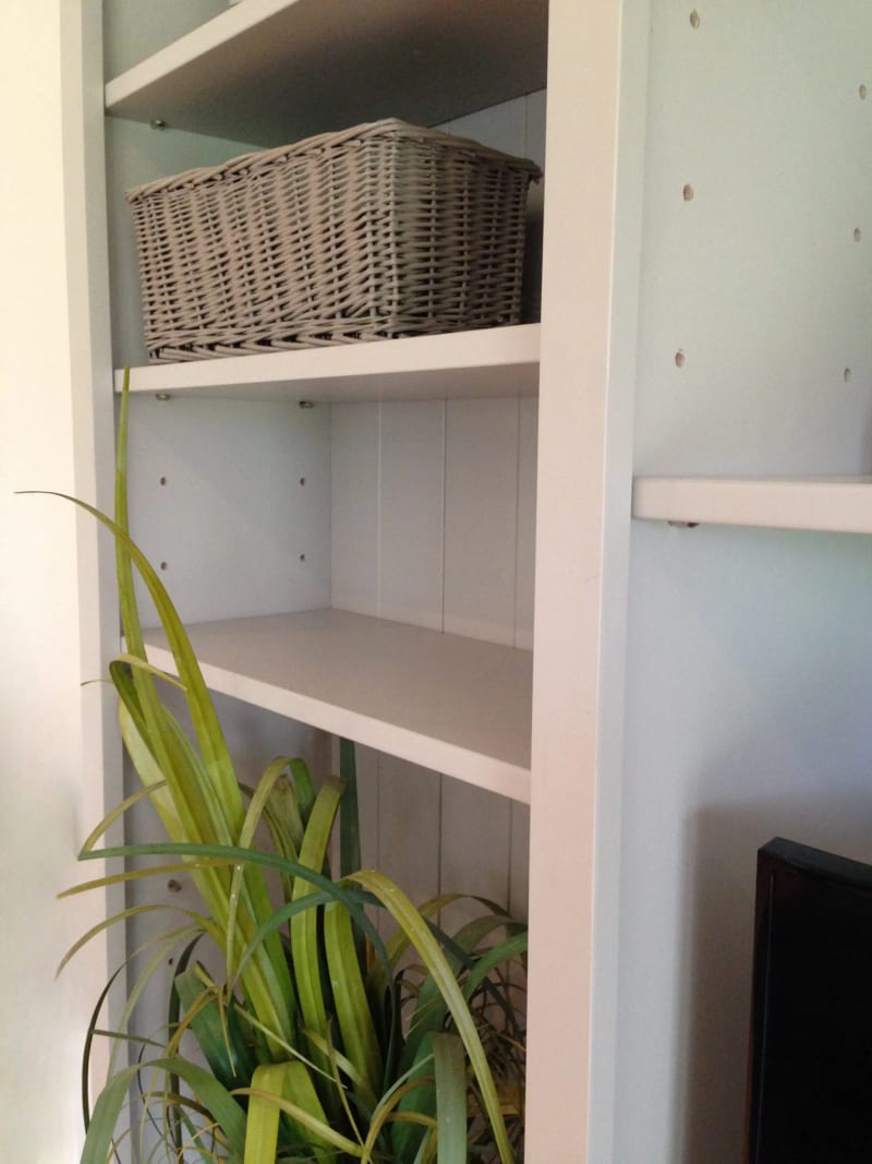 Close view of an outer shelved bay showing a grey wicker basket, adjustable shelves with metal pins, the painted tongue-and-groove back boards and a spider plant in the foreground