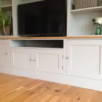 Angled view of the cupboard base showing four shaker doors with chrome knobs, the open central television recess beneath the oak top and the wide plinth on oak floorboards