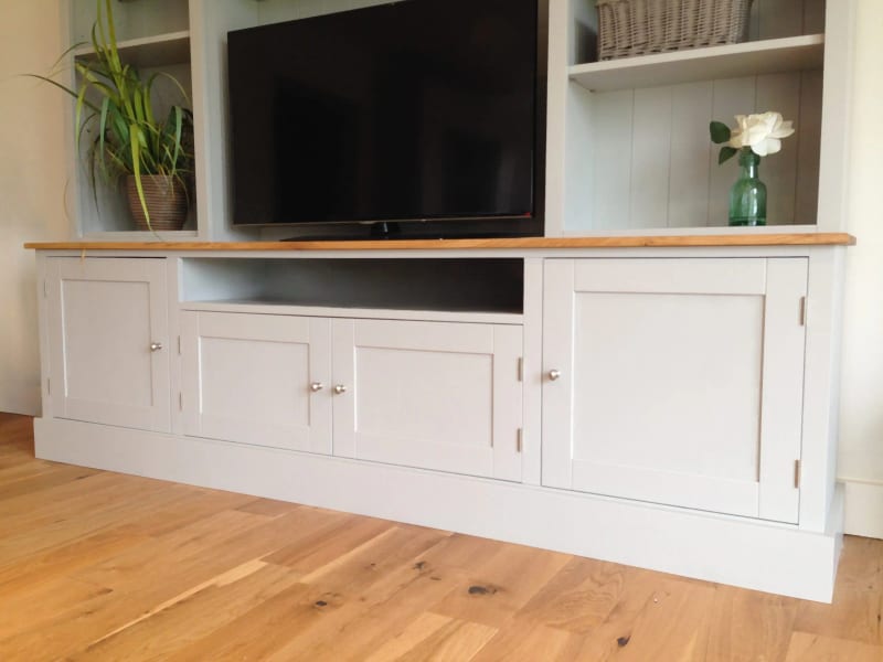 Angled view of the cupboard base showing four shaker doors with chrome knobs, the open central television recess beneath the oak top and the wide plinth on oak floorboards