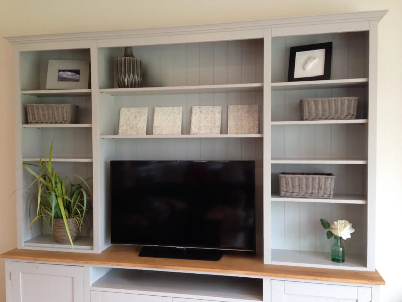 Front view of the upper bookcase section showing three bays of adjustable shelves, wicker baskets, framed prints, a ribbed vase, four small canvases and a television on the oak top