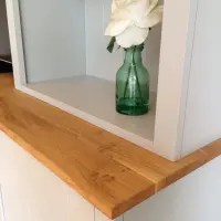Close-up of the corner of the oak top where it meets the bookcase upright, showing the grain and sawn edge beside a white rose in a green glass bottle on the shelf above
