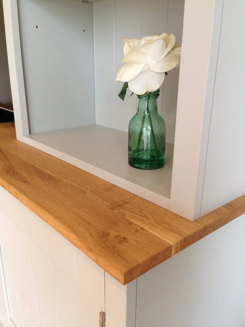 Close-up of the corner of the oak top where it meets the bookcase upright, showing the grain and sawn edge beside a white rose in a green glass bottle on the shelf above