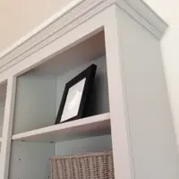 Looking up at the top corner of the painted bookcase, showing the moulded cornice, a black framed print and a grey wicker basket on the shelves against the tongue-and-groove back