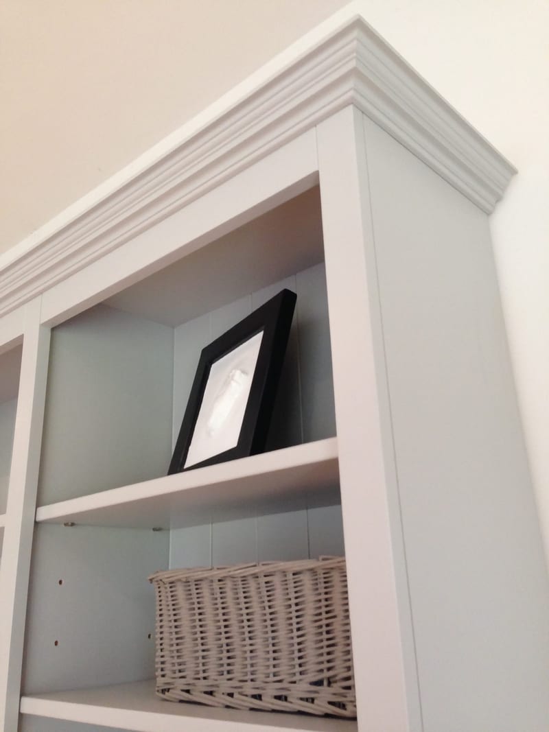 Looking up at the top corner of the painted bookcase, showing the moulded cornice, a black framed print and a grey wicker basket on the shelves against the tongue-and-groove back