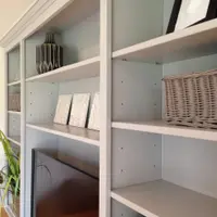 Angled view along the shelves from the side, showing the adjustable shelves with pin holes, wicker baskets, a ribbed grey vase, three small canvases and the top edge of the screen
