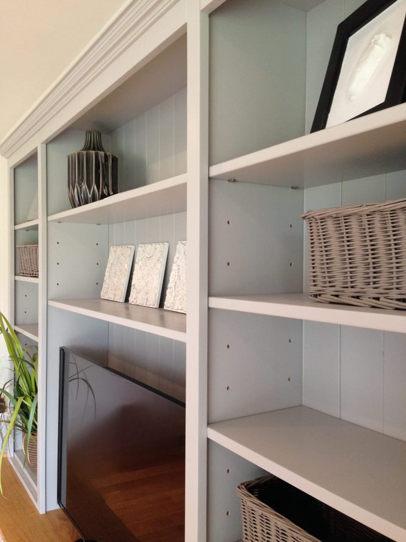 Angled view along the shelves from the side, showing the adjustable shelves with pin holes, wicker baskets, a ribbed grey vase, three small canvases and the top edge of the screen