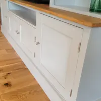 Low angled view of the cupboard base showing the shaker doors with chrome knobs and hinges, the oak worktop overhang, the open television recess and the plinth on oak floorboards