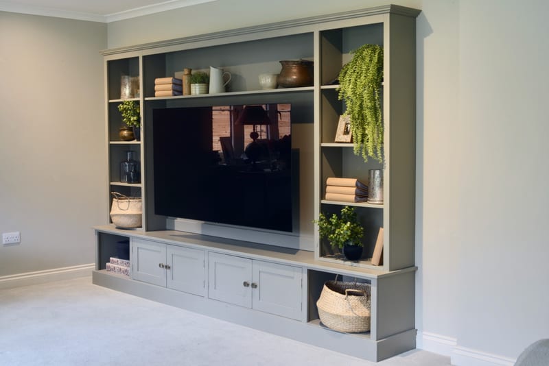 Three-quarter view of the grey-green painted media wall unit against a pale stone wall, with four shaker cupboard doors, pale ceramic knobs, open side shelving and a television in the central recess