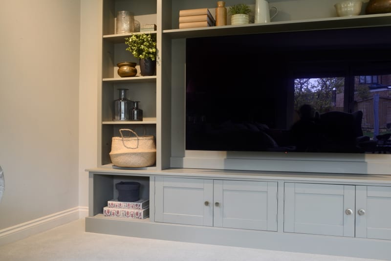 Close view of the left end of the painted media unit, showing an open shelf column with a plant, brass pot and woven basket above a low cubby holding patterned boxes and a navy tub
