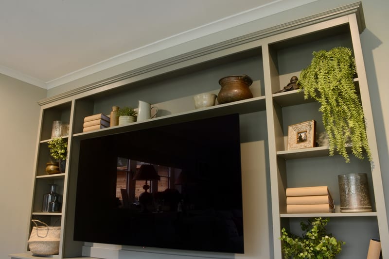 Upward view of the top of the painted media unit, showing the moulded cornice, ceiling coving and upper shelves holding a copper pot, a white jug, a cream bowl, books and a long trailing plant
